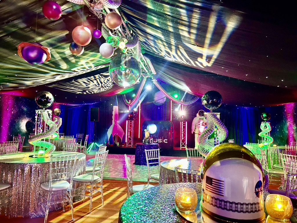 Colorful decorated event room with metallic tables, chairs, hanging ornaments, and space-themed decorations including a NASA helmet, spiral structures, glittery curtains, and multicolored lighting.