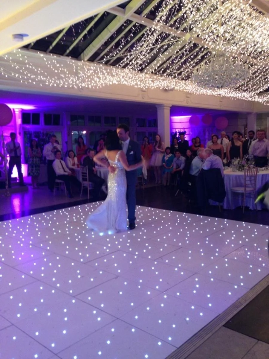 Couple dancing on a lit dance floor at a wedding reception, with guests watching under decorated ceiling with string lights and purple ambient lighting.