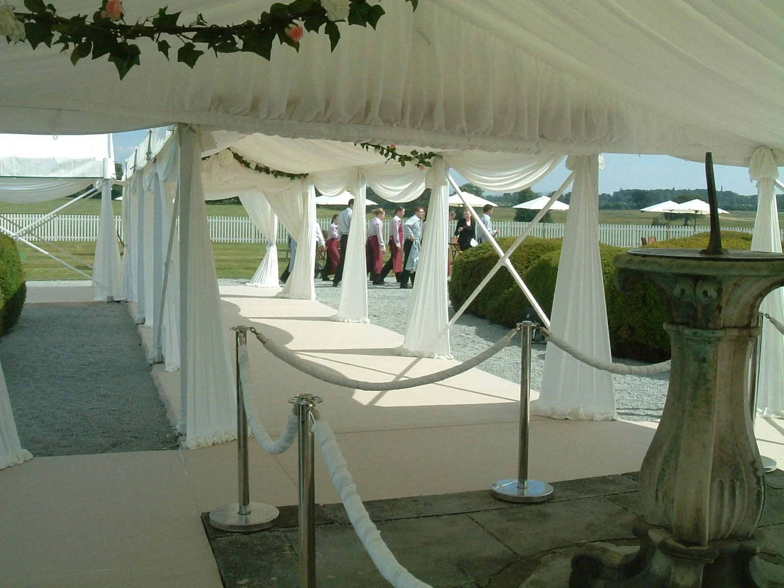 White wedding ceremony aisle under a decorated canopy with a group of waitstaff or servers walking in the background outdoors.