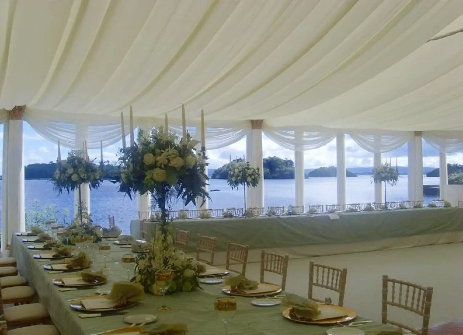 A decorated outdoor banquet area by a large body of water, with a white tent, elegant floral arrangements, and dining tables set for an event.