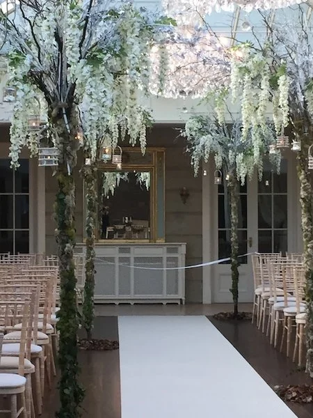 Indoor wedding aisle decorated with hanging white flowers and greenery, with chairs on both sides, and a white carpet.