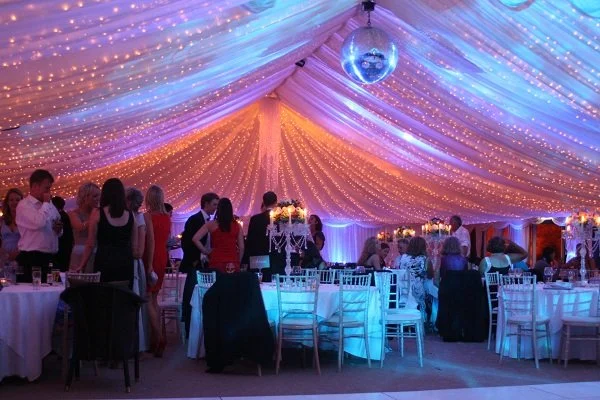 People gathering inside a decorated event tent with draped fabric, string lights, and a disco ball at a formal celebration or wedding reception.