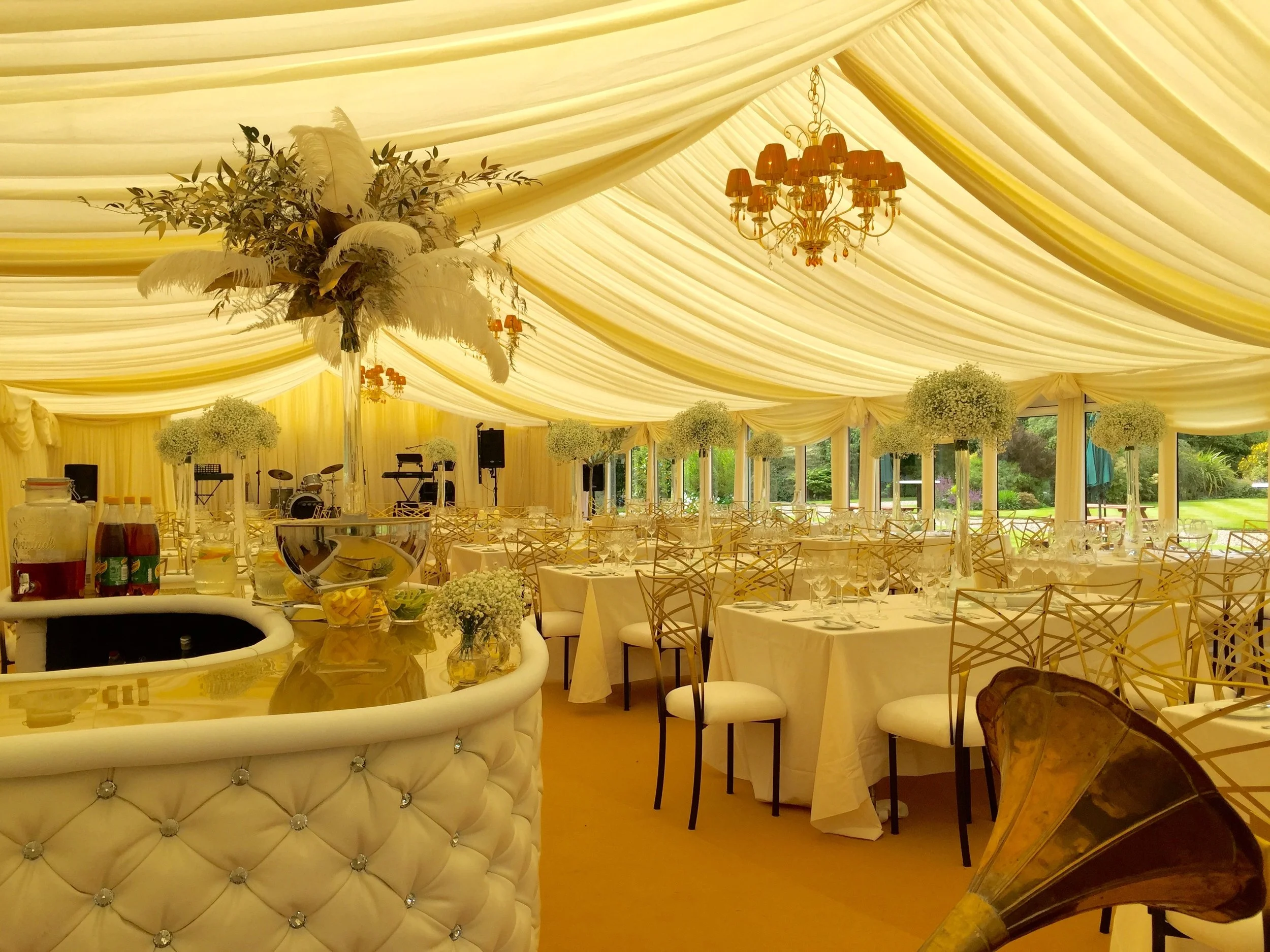 Elegant event space with cream-colored draped ceiling, ornate chandelier, round tables with white tablecloths, floral centerpieces, and gold chairs. Open outdoor view with greenery visible outside.