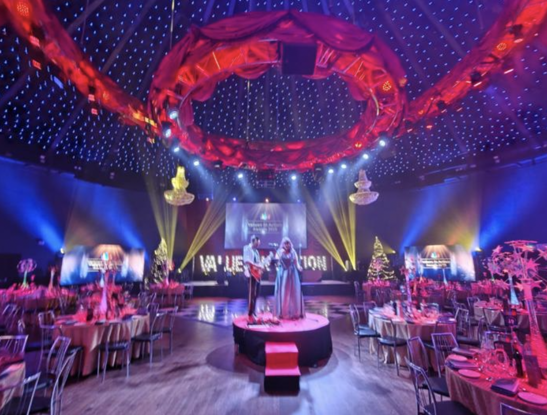 This image captures a breathtaking crystal chandelier suspended like a glowing jewel beneath a canopy of rich, draped fabric, bathing the space in violet and blue light.