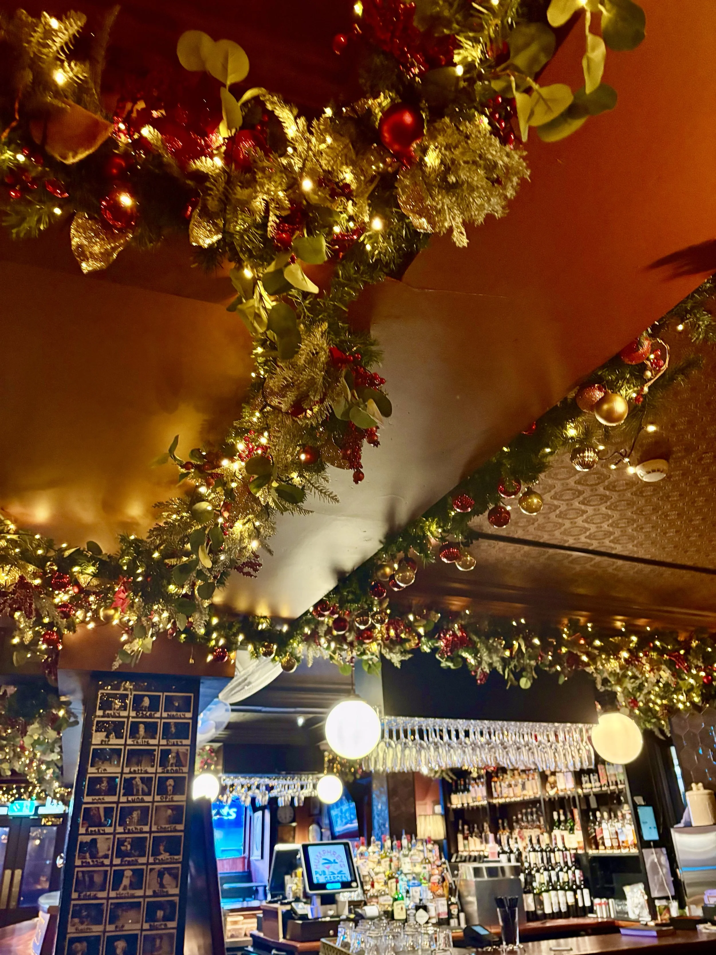Decorative Christmas garland with red and gold ornaments hanging over a bar area in a restaurant or pub, with various bottles and glasses on the shelves behind.