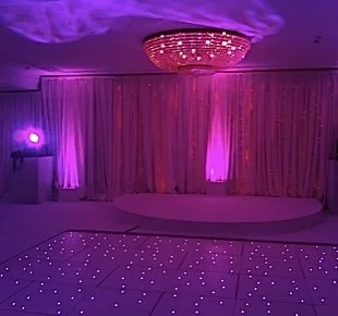 An event venue with purple lighting, a chandelier on the ceiling, a stage with curtains, and a dance floor with illuminated dots.