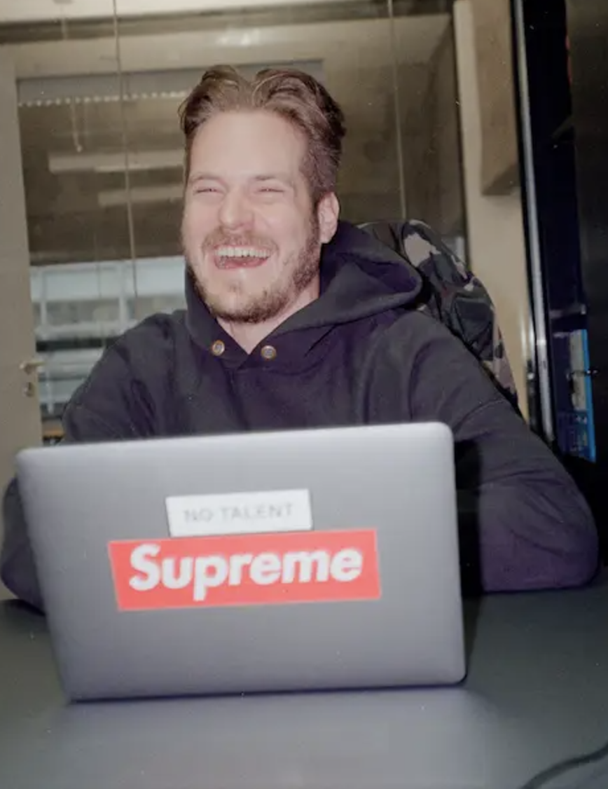 Alexander de Lukowicz, sitzt an einem Tisch mit einem Laptop, der Aufkleber mit den Marken "Supreme" und "NO TALENT" hat, im Hintergrund ein moderner Raum mit Glaswänden.
