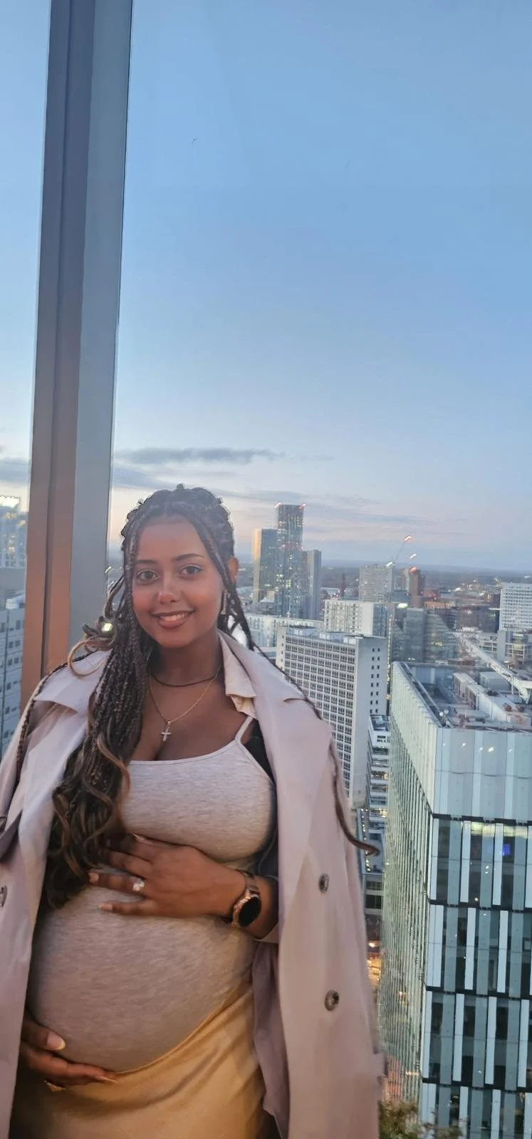A pregnant woman with long braids, smiling, standing inside a high-rise building with a city view of tall buildings and skyscrapers during sunset.