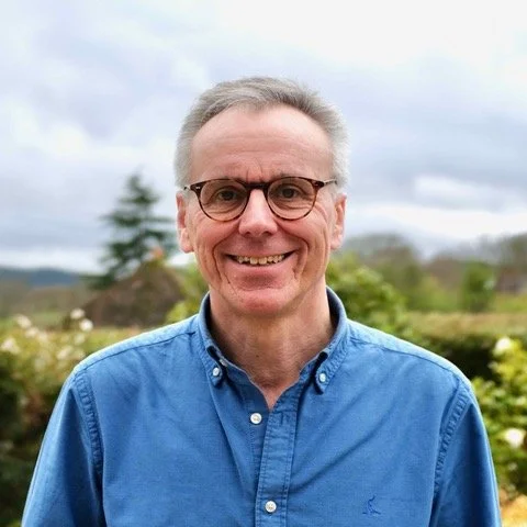 Smiling middle-aged man wearing glasses and a blue button-up shirt outdoors with trees and cloudy sky in background.