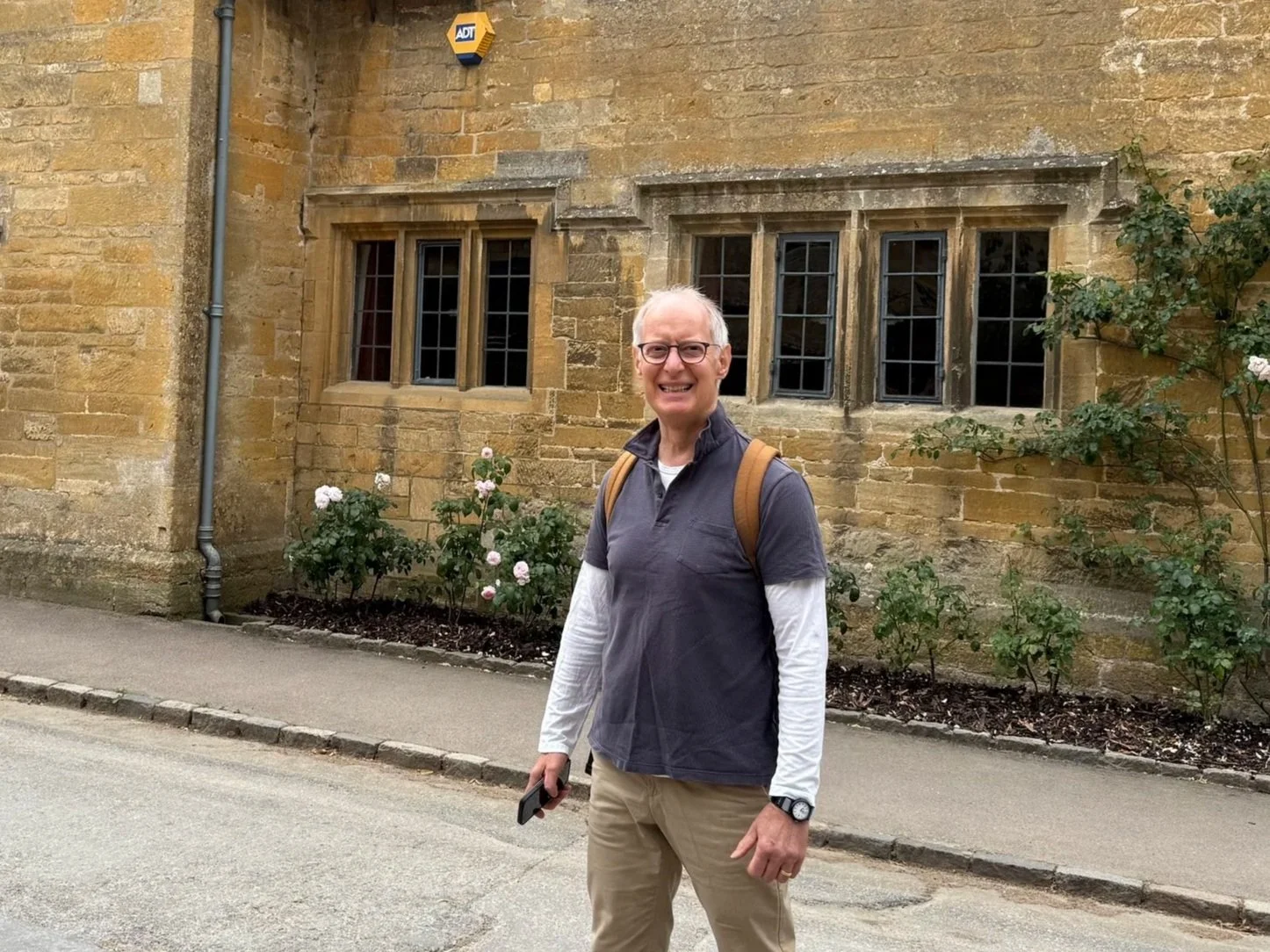 An elderly man with glasses, wearing a gray short-sleeve shirt over a white long-sleeve shirt and khaki pants, stands on a sidewalk in front of a historic brick building with flowers and shrubs. He is smiling, holding a phone, and wearing a backpack.