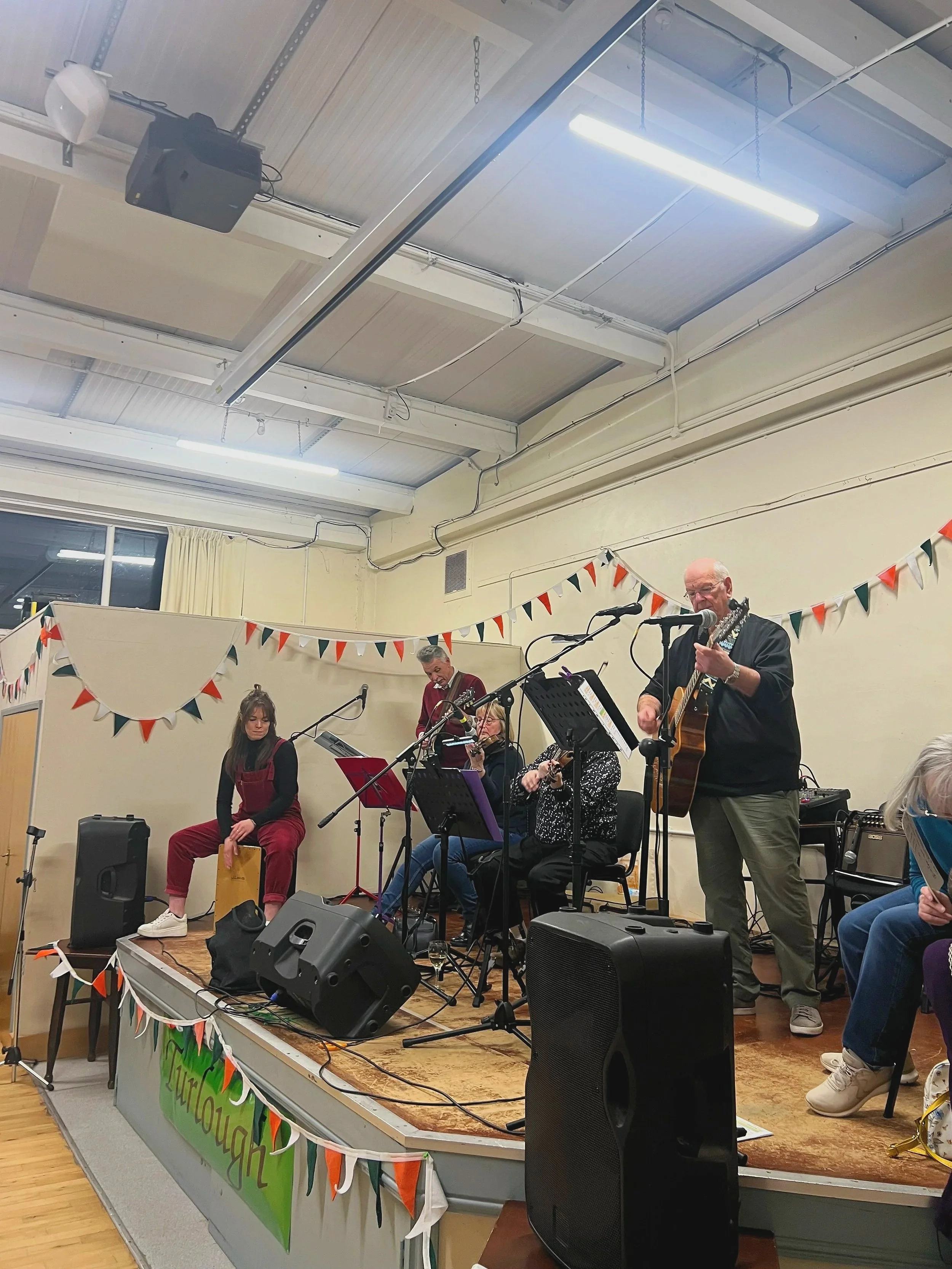 A group of musicians performing on a stage in a decorated room with bunting. One man is playing an acoustic guitar and singing into a microphone, while three women are playing instruments or singing. A banner with text and colorful decorations is hung on the front of the stage.