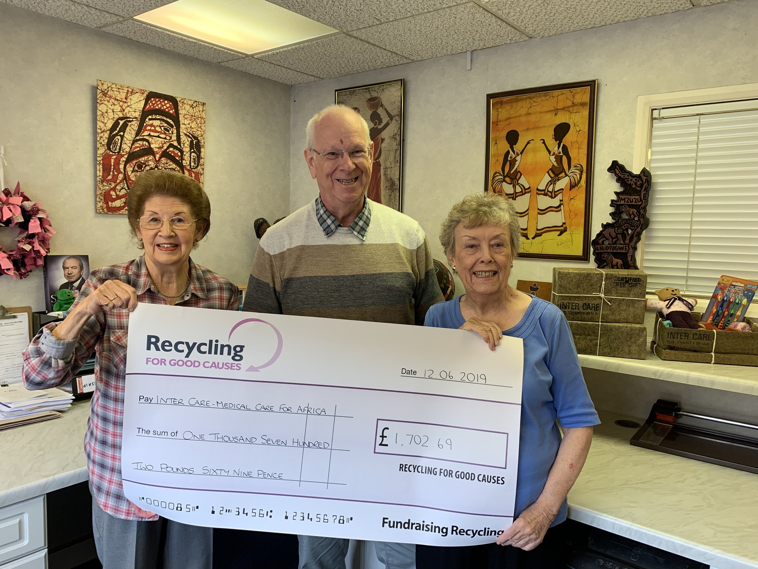 Three people standing behind a large check for £1,702.69 made out to the charity Inter Care. The woman on the left is holding the check and smiling, the man in the middle is smiling, and the woman on the right is also smiling.