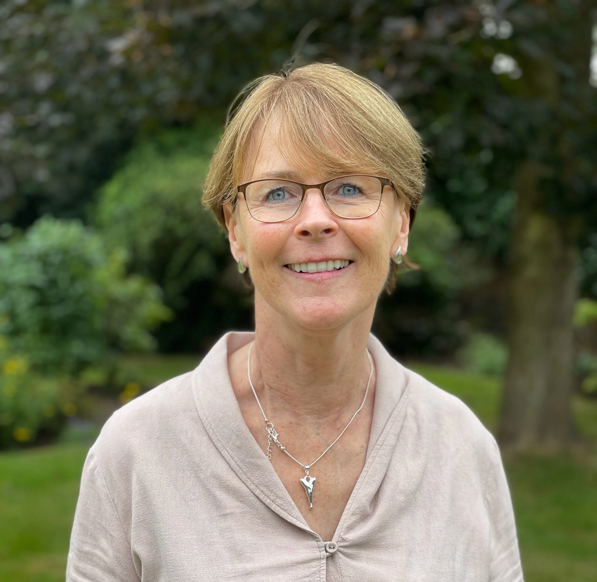 Close-up of a smiling middle-aged woman with short hair, glasses, wearing a beige cardigan, outdoors with blurred green foliage in the background.