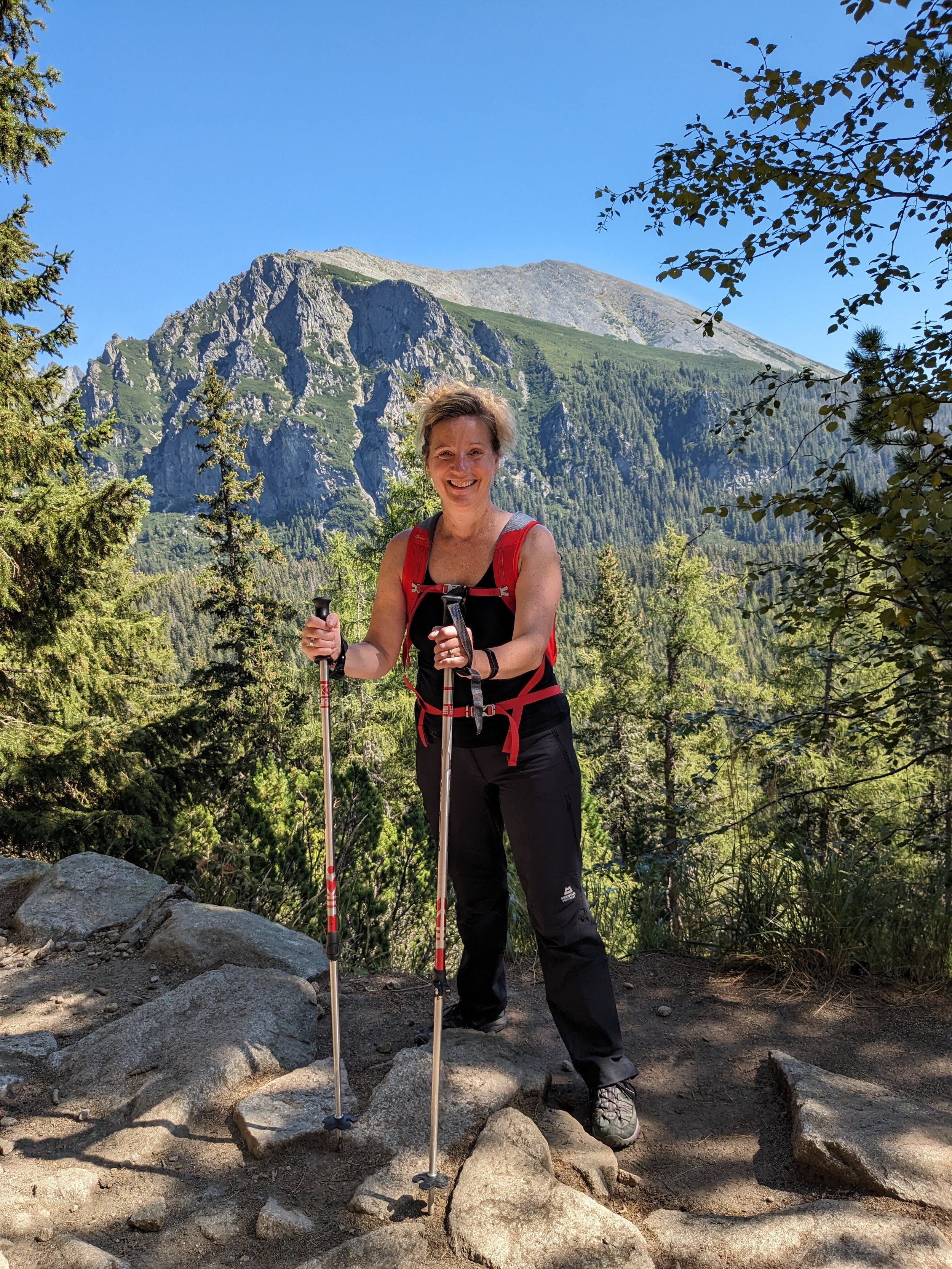 Woman hiking outdoors in a mountainous forest, smiling and holding trekking poles, with a large mountain in the background.
