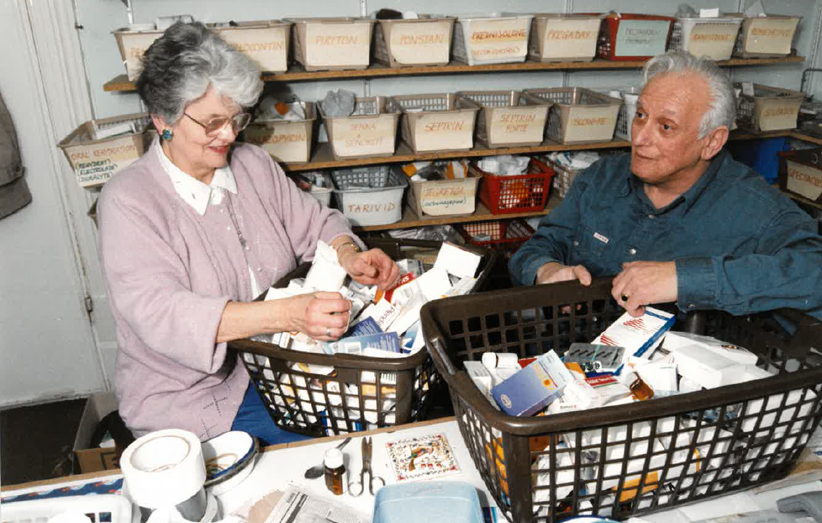 The founders of Inter Care: A woman and man sorting medical supplies or medications at a table in a storage room, with labeled bins on shelves behind them.
