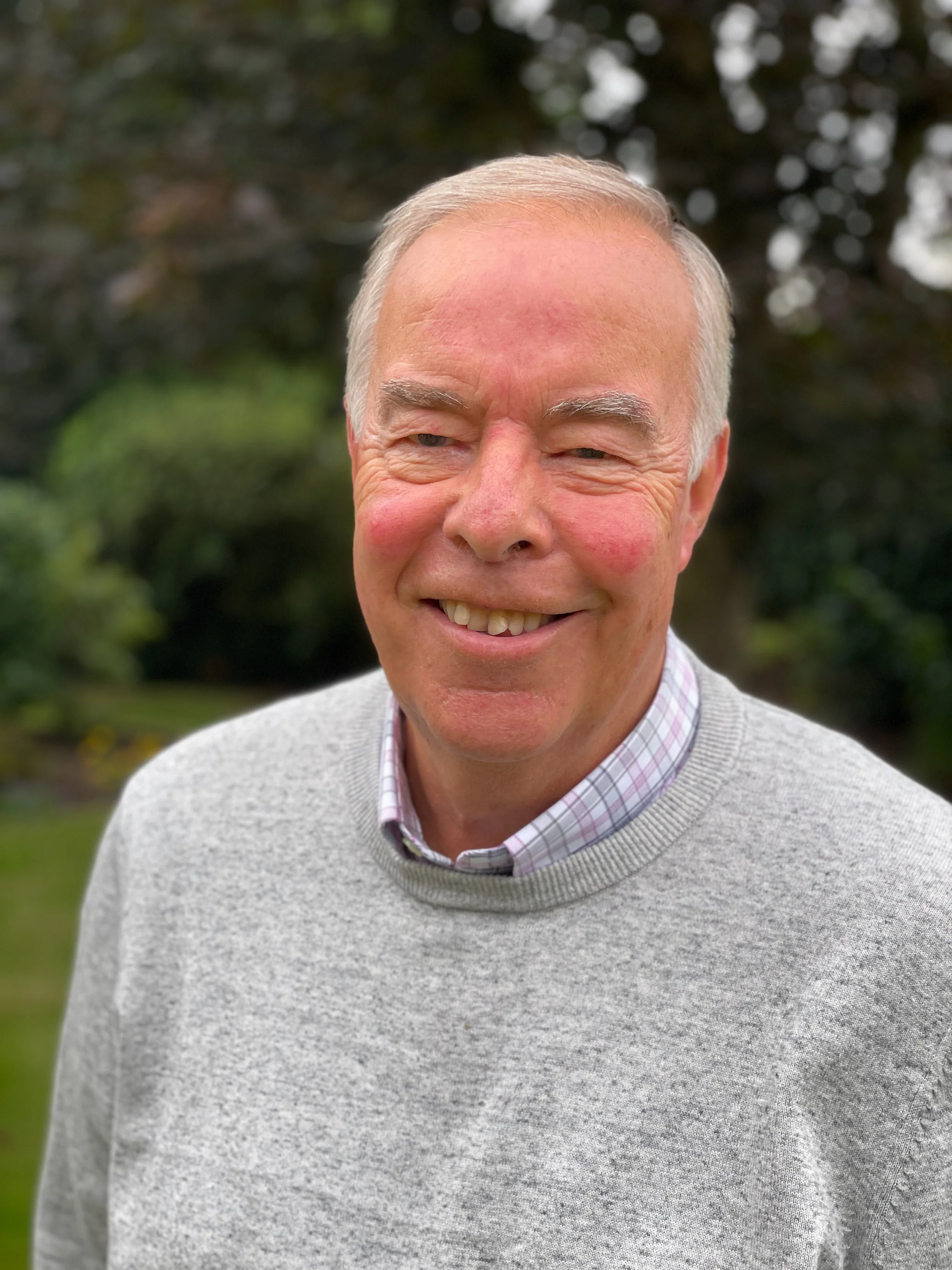 Close-up of an elderly man smiling outdoors, wearing a light gray sweater over a collared shirt with a checkered pattern, with a blurred background of trees and greenery.