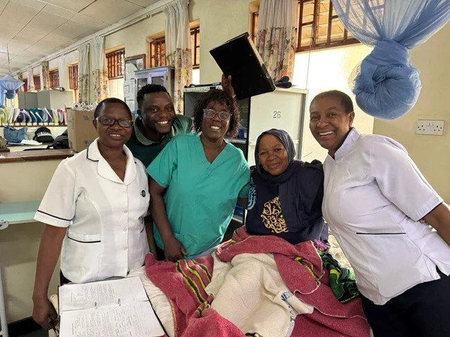 Five women nurses are gathered around a patient lying in bed, smiling. The patient is covered with pink, red, and white blankets, and is receiving medical care. The room has large windows with curtains in the background.