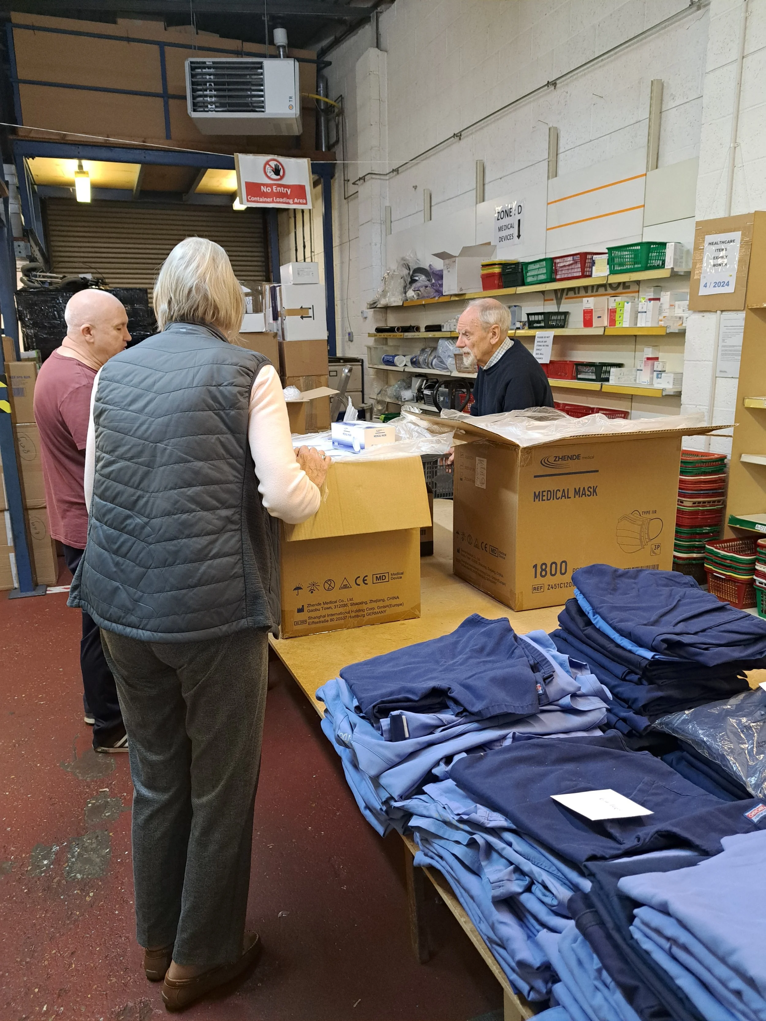 A woman and a man talking with a worker behind a table filled with folded clothing in a warehouse. Shelves with boxes and supplies are in the background.