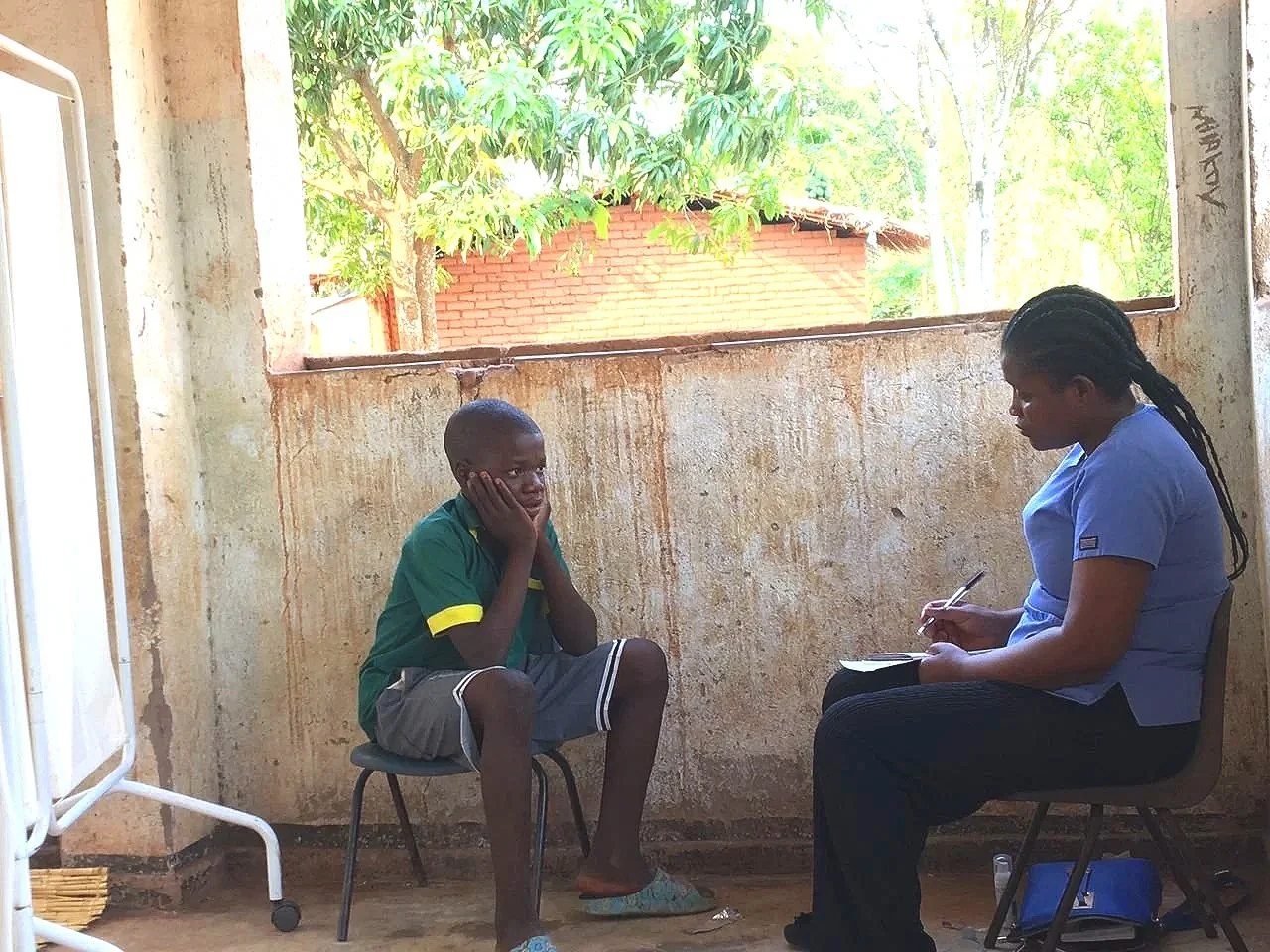 A young boy sitting on a small stool with his hands on his cheeks, talking to a female healthcare professional or counselor who is taking notes, in a room with unfinished, weathered walls and a large window showing greenery outside.