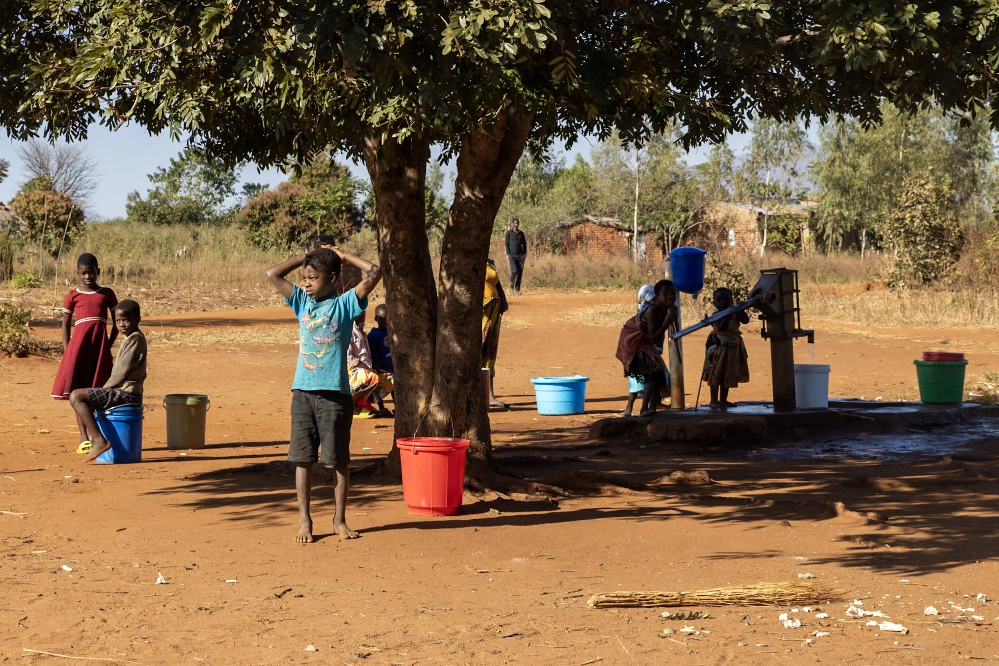 Children in a rural area in Africa around a large tree, some sitting on buckets and others standing, with water pumps and buckets nearby, and a person walking in the background.