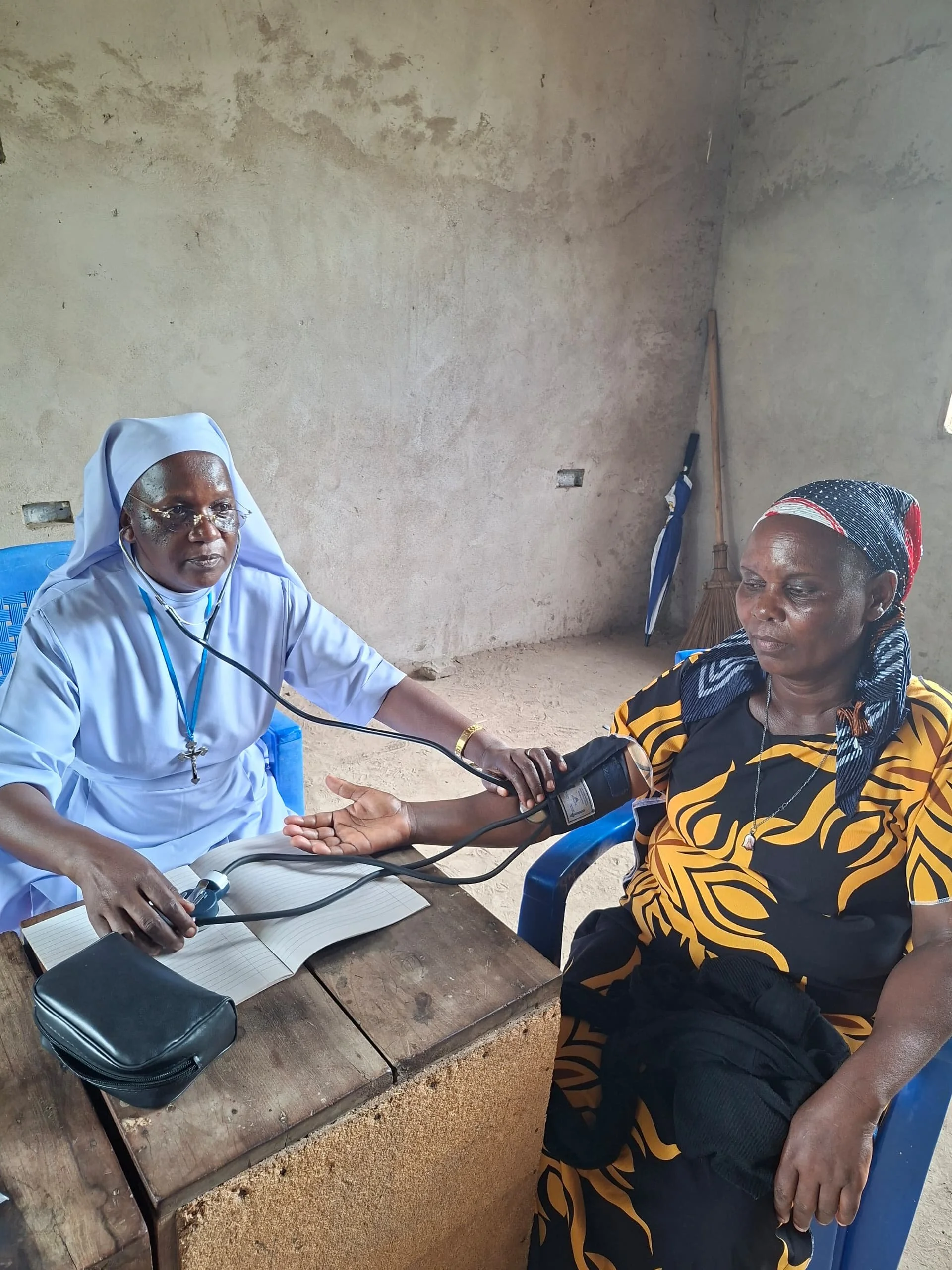 A healthcare worker, dressed in a light blue uniform and wearing glasses, is taking the blood pressure of a woman seated on a blue plastic chair. The woman is wearing a black and yellow patterned dress and a headscarf. They are seated indoors against a plain, unfinished wall, with a broom and an umbrella in the background.