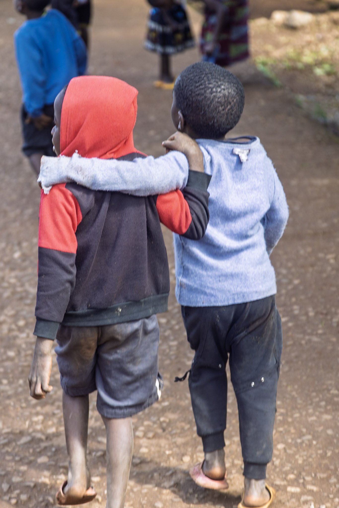 Two children walk side by side outdoors, one wearing a red hooded sweatshirt with a black backpack, and the other wearing a light blue shirt. The child in blue has their arm around the shoulder of the child in red. They are walking on a dirt path, with other children in the background.