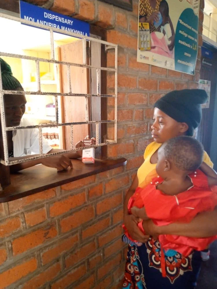 A woman holding a child stands at a dispensary window counter with a brick wall background, speaking to a healthcare worker.