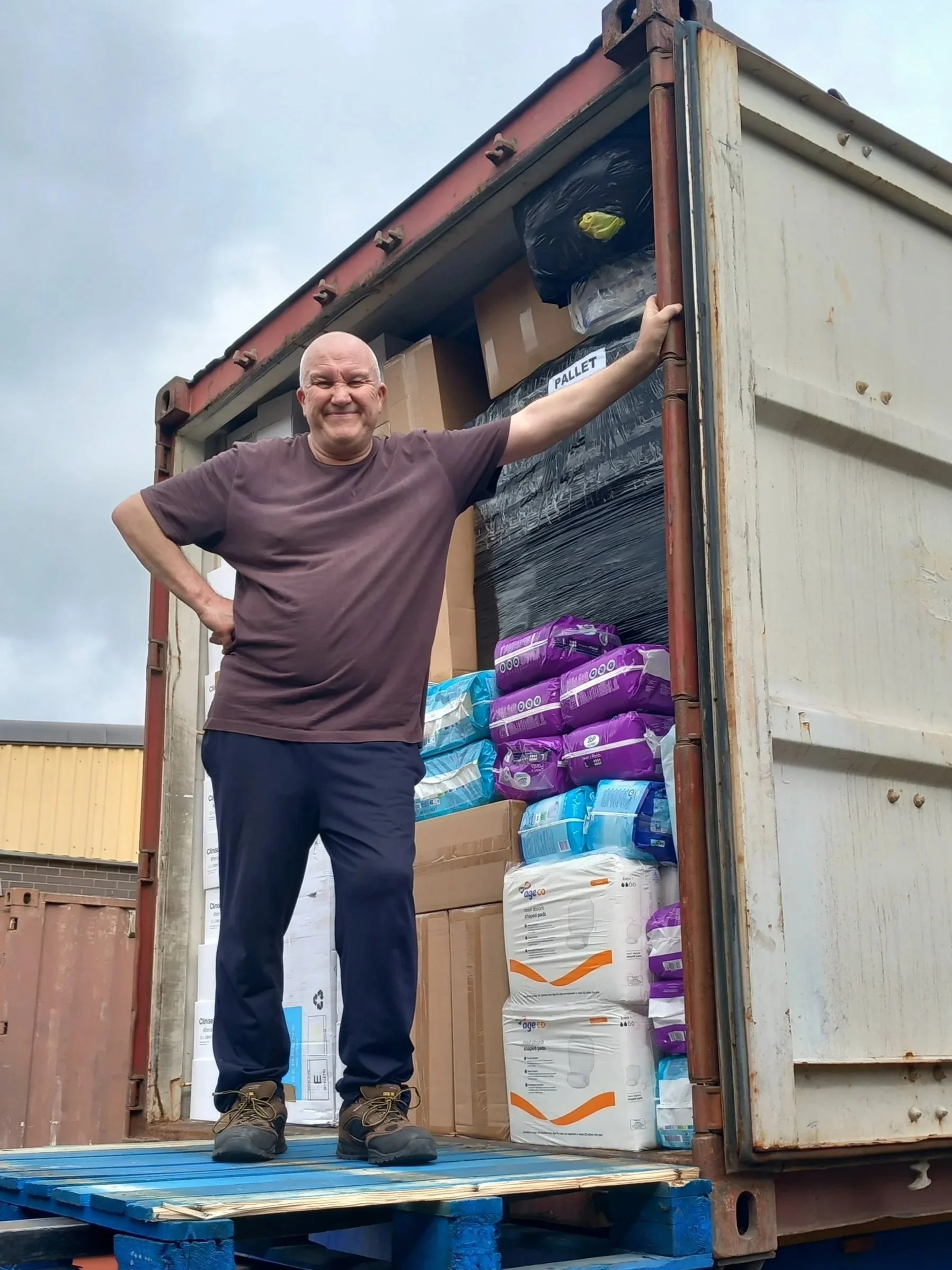 Smiling man standing at the open back of a shipping container filled with various boxes and bags, outdoors under a cloudy sky.