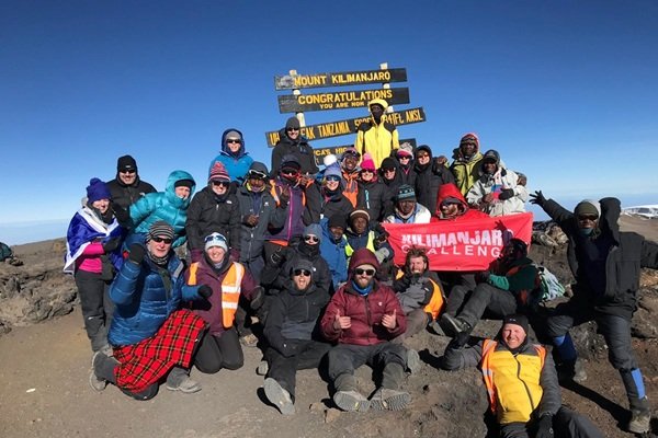 Group of hikers at Mount Kilimanjaro summit, holding a red banner and posing for a photo near signage.