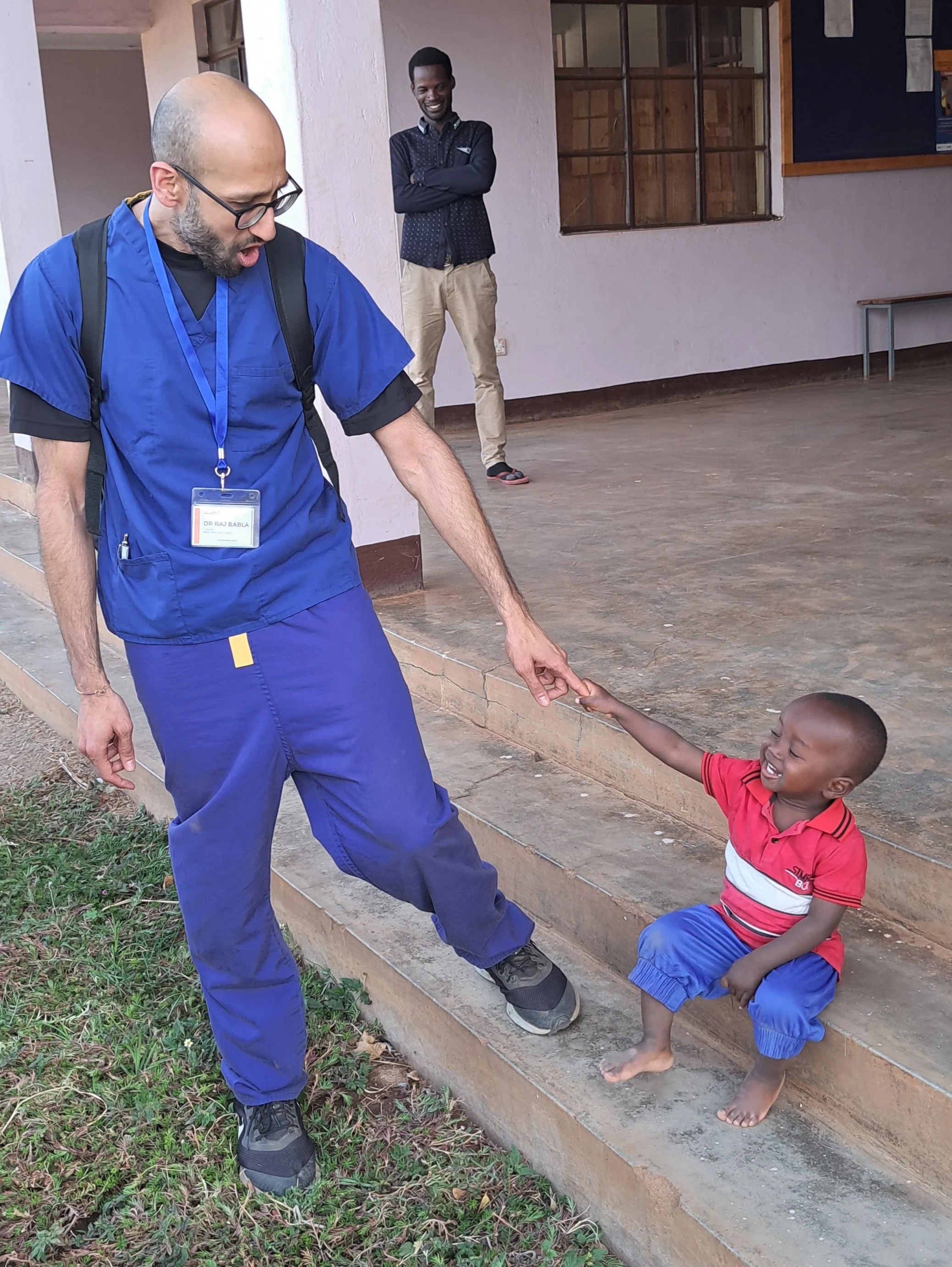 A man dressed in blue medical scrubs with a backpack and glasses is reaching down to hold the hand of a smiling young boy in a red and blue striped shirt and blue shorts, who is standing barefoot on a concrete step. Another man in the background, wearing a dark shirt and beige pants, is standing with arms crossed and smiling.