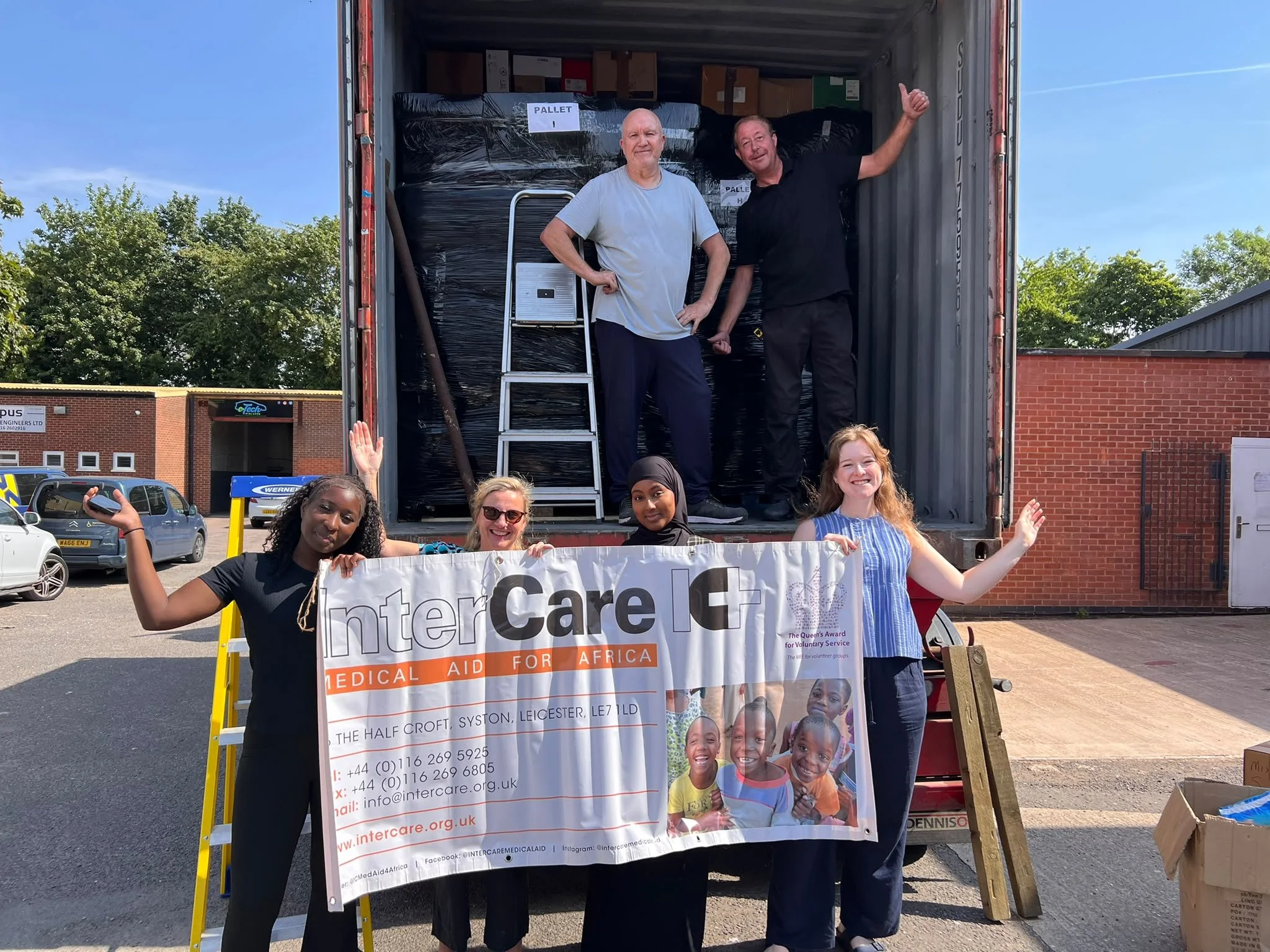 A group of Inter Care's Staff standing behind a large banner for Inter Care - Medical Aid for Africa, with a container loaded with supplies behind them on a sunny day.