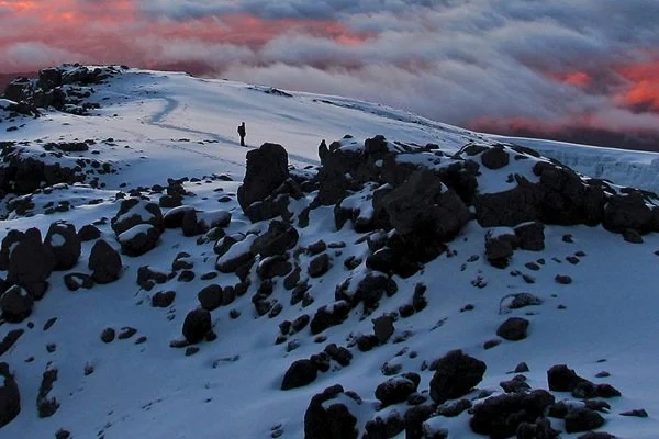 A person walking along a snowy landscape with rocks and hills, under a sunset sky with clouds.