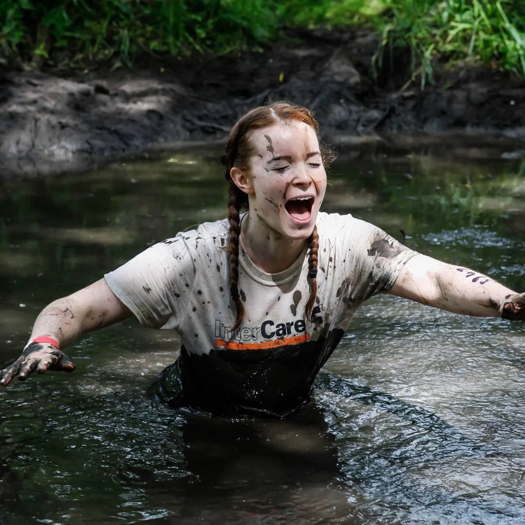 A woman with braided hair, covered in mud, wading in a muddy creek, with her arms outstretched and a joyful expression taking part in an Inter Care fun-run