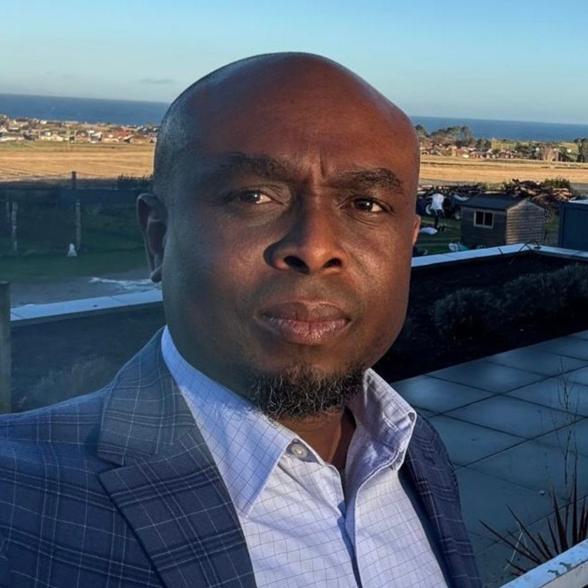 A man with dark skin, a bald head, and a beard, dressed in a blue checked suit and light blue shirt, standing outdoors with an ocean, cityscape, and open fields in the background.