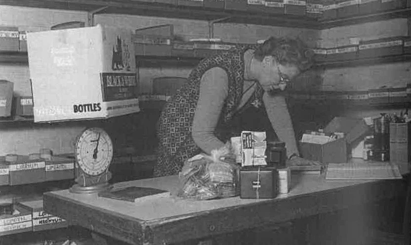 Two senior adults, a woman and a man, sorting medications into baskets in a pharmacy or medical storage room with labeled shelves in the background.
