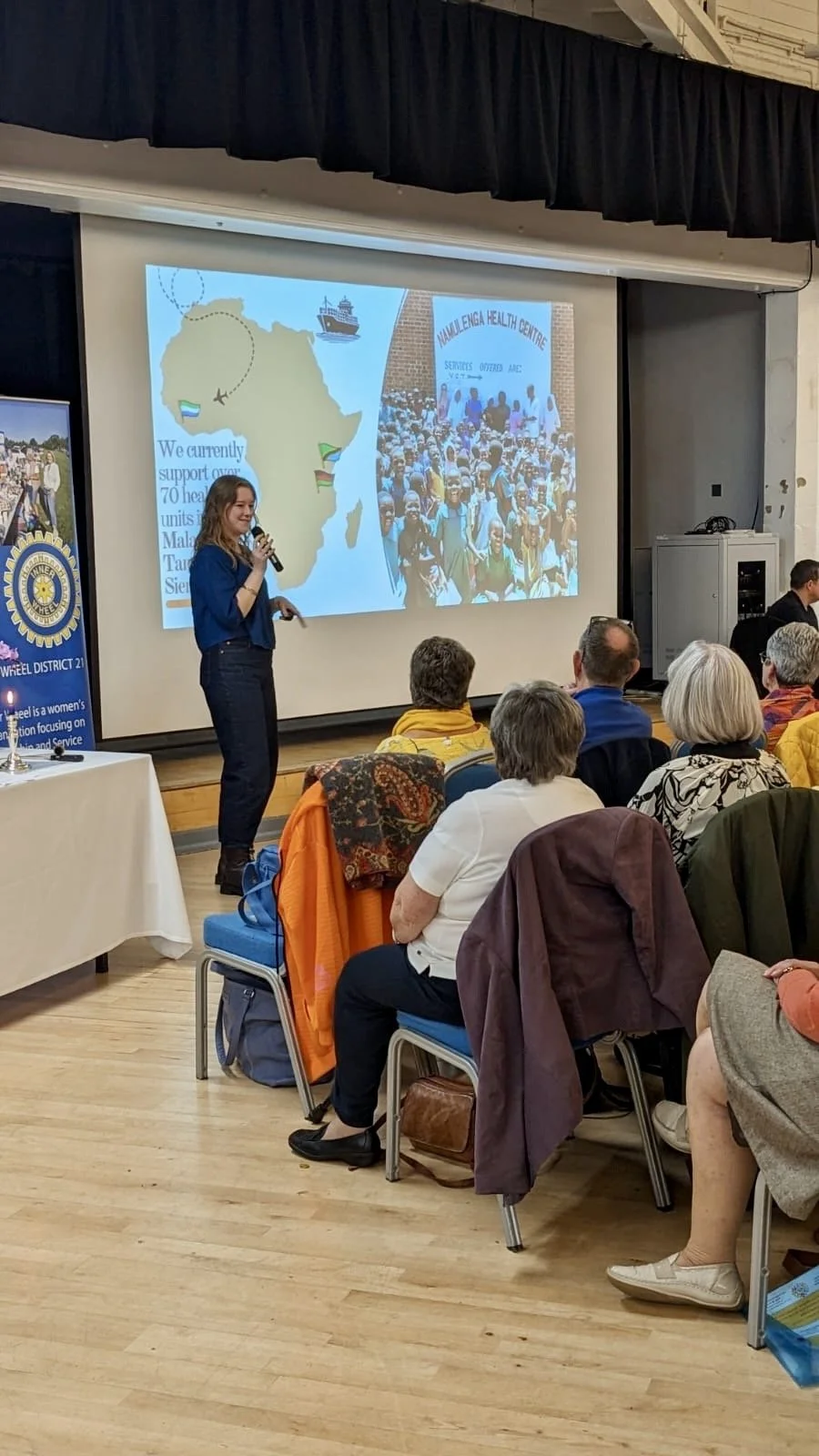 A woman giving a presentation in front of an audience in a conference room, with a slide showing a map of Africa and a photo of a large group of people at the Health Centre