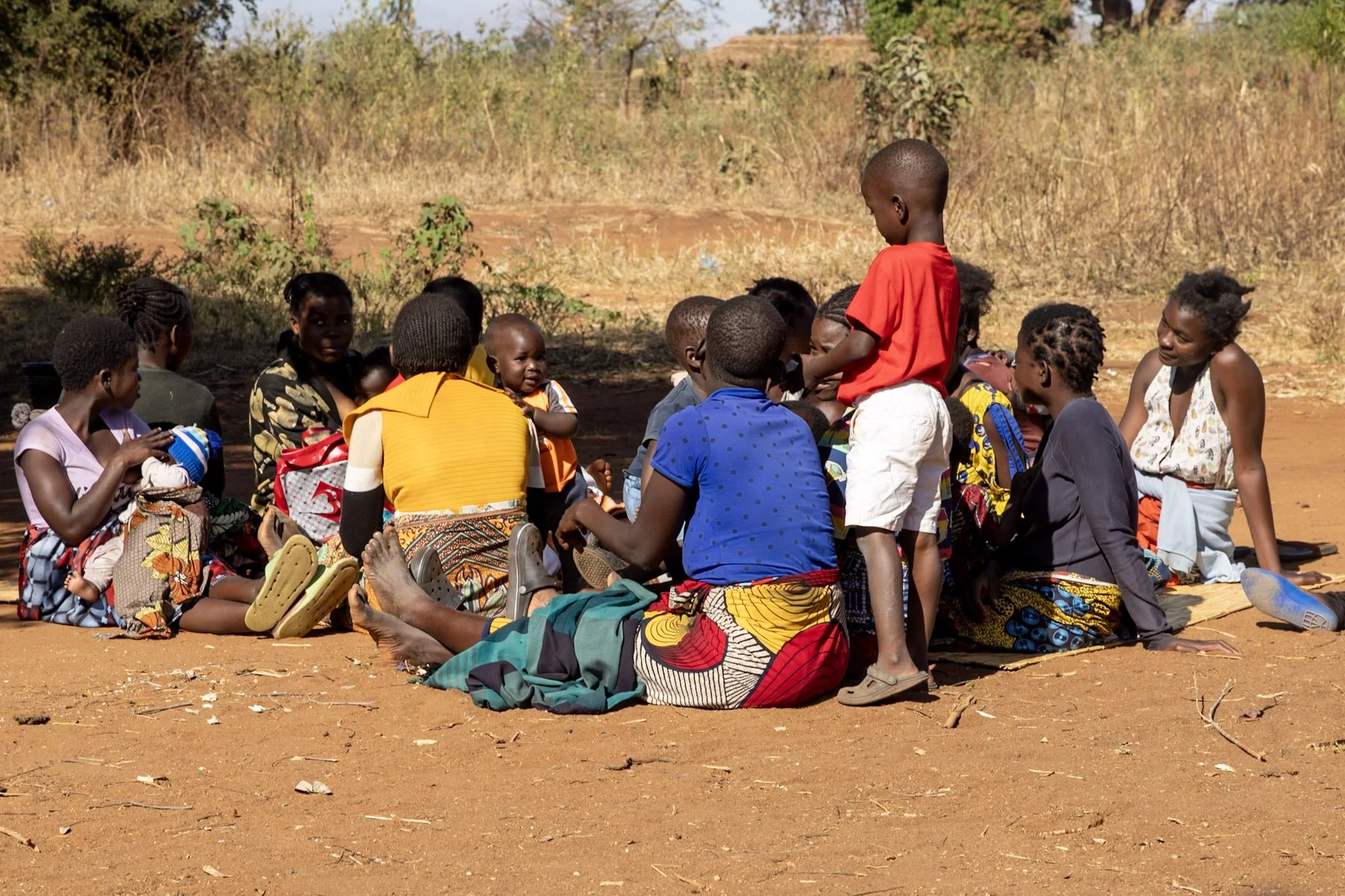 Group of children and young adults sitting on the ground outdoors, some engaging in conversation. The scene takes place in a dry, rural area with sparse vegetation and a dirt ground.