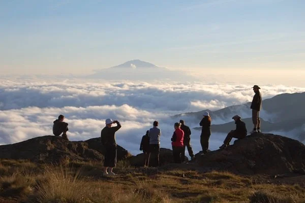 Group of people standing and sitting on rocky terrain on a mountain, overlooking a sea of clouds with a mountain in the distance.