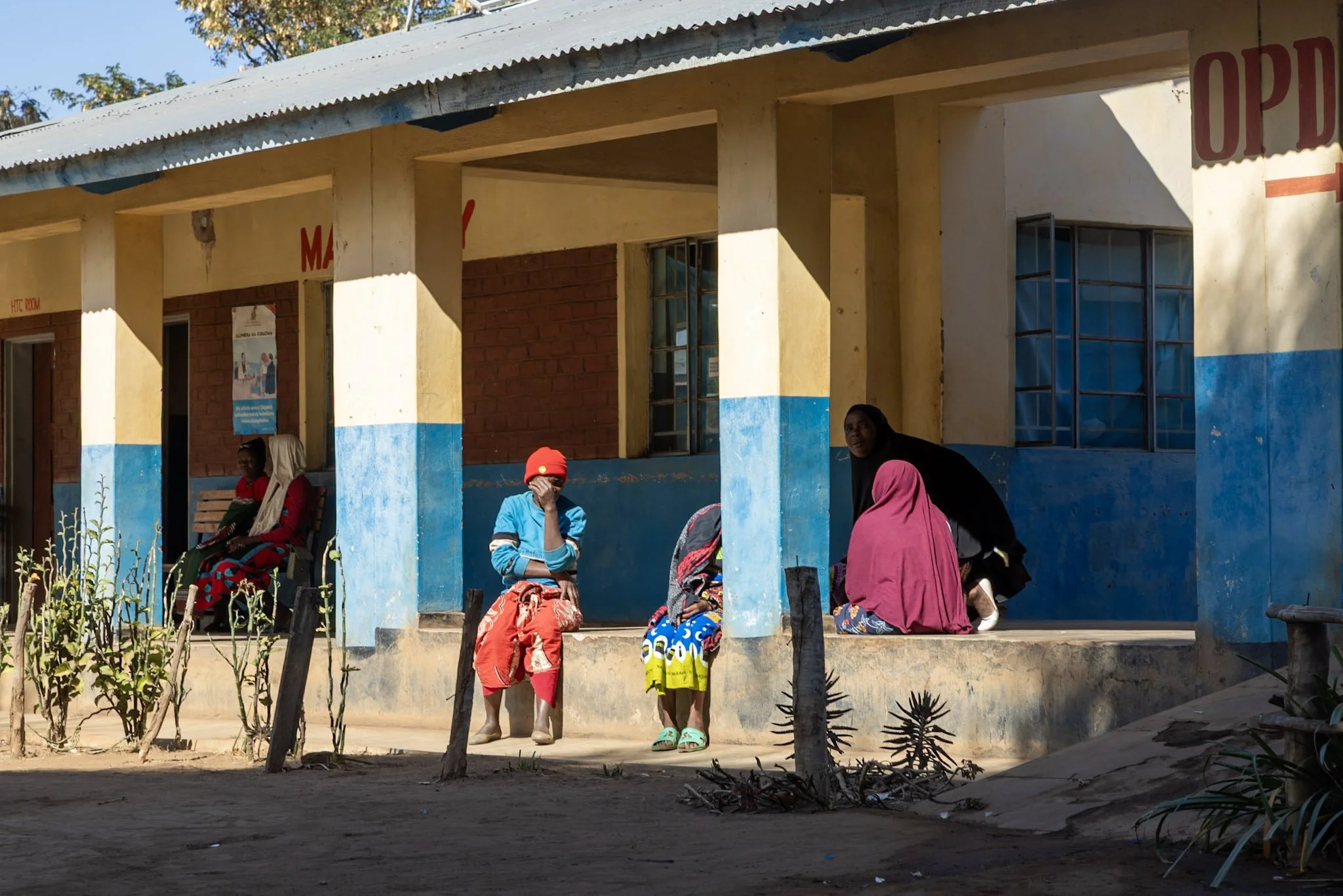A group of women and children sitting and standing outside a yellow and blue building with a corrugated metal roof. Some women are seated on chairs, others are standing or crouching. The building has large windows with metal bars and a sign partially visible.
