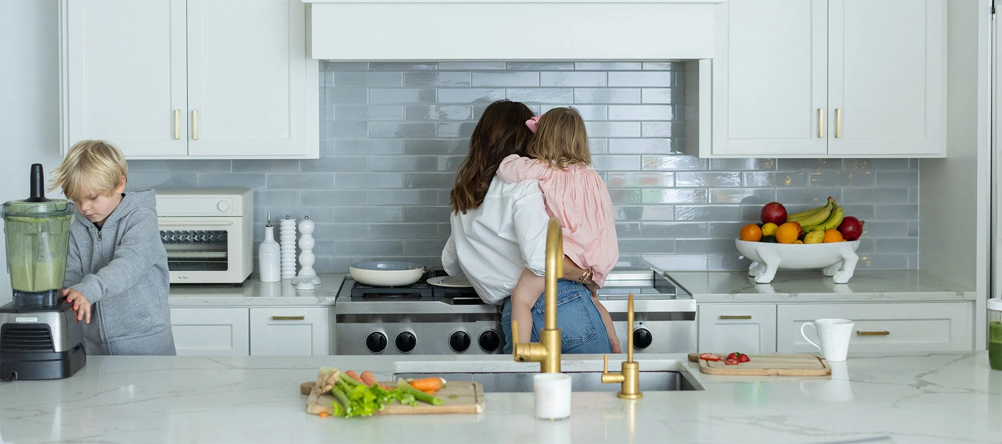 Three children in a modern kitchen, with one girl hugging a younger girl, another boy blending vegetables in a blender on the counter.