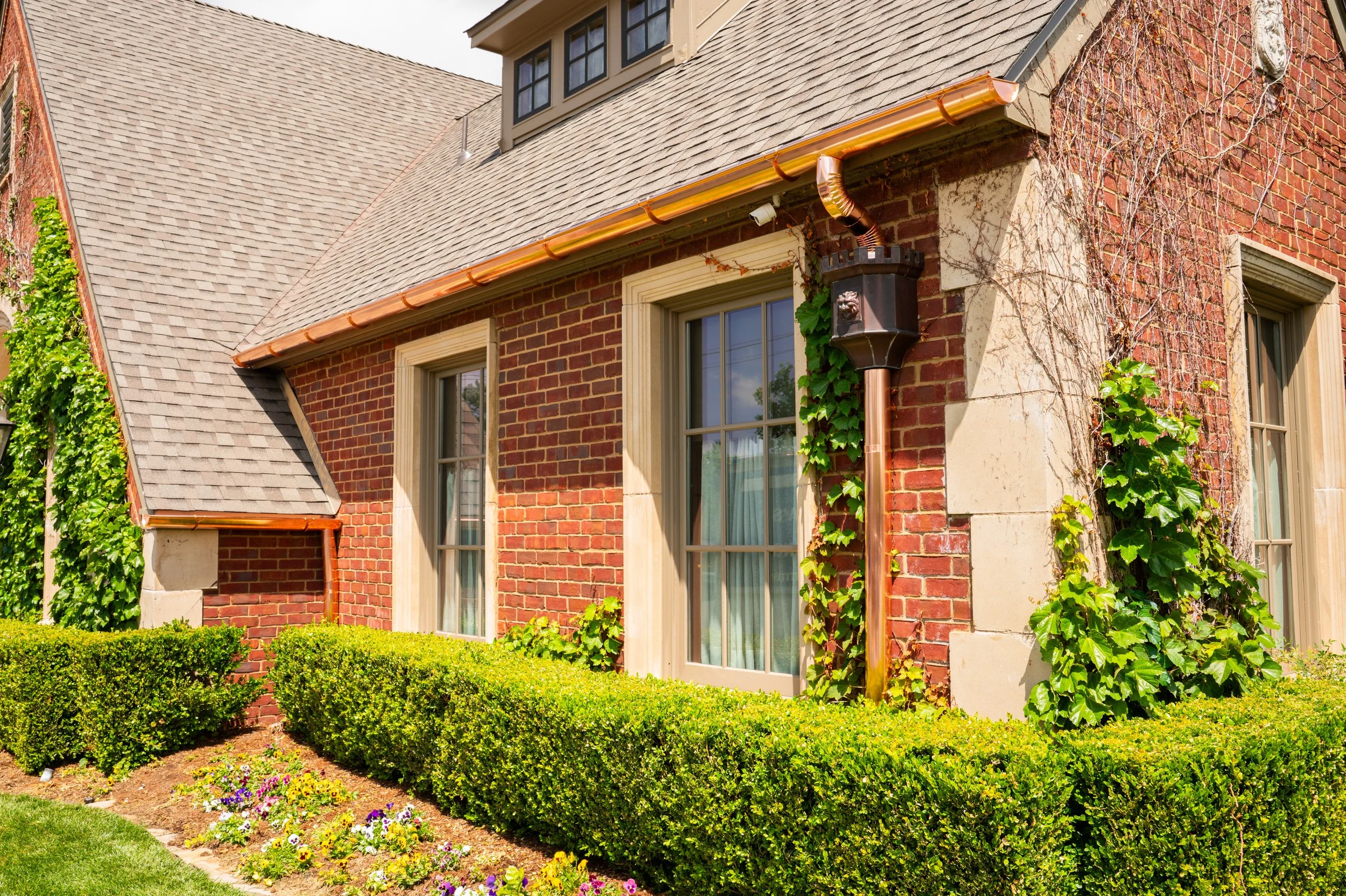 A red brick house with white trim and large windows, surrounded by a well-maintained lawn with a flower bed and trimmed bushes, and a copper gutter system.
