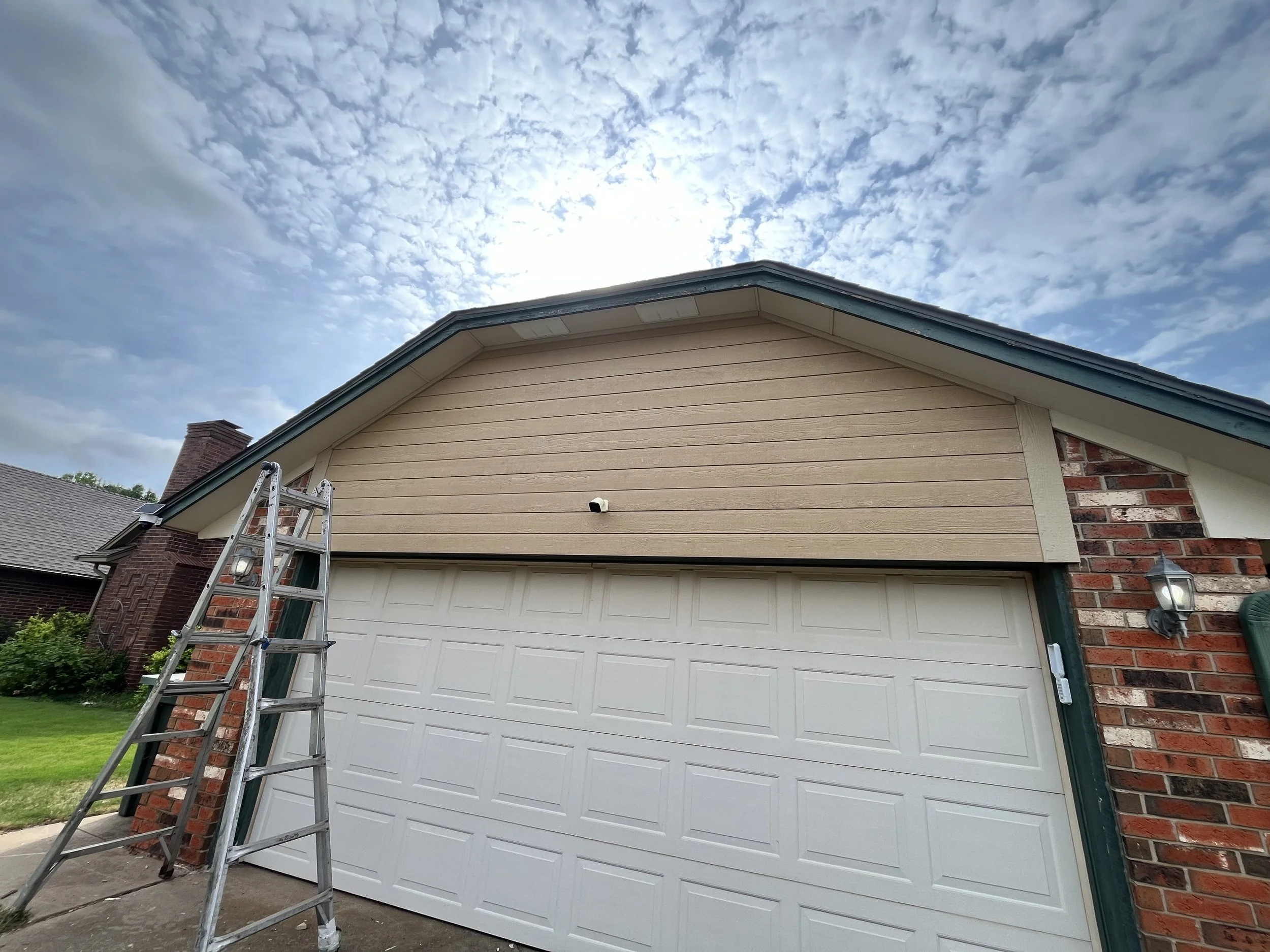 View of a house with a beige wooden siding and a white garage door, a ladder leaning against the house, partly cloudy sky above, and a brick chimney on neighboring house.