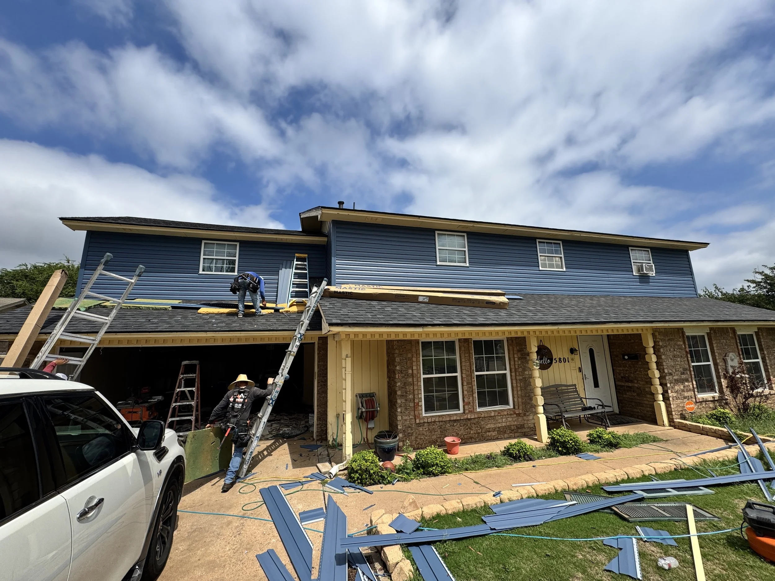 Two workers on a ladder installing blue siding on the upper level of a two-story house. The house has a brick lower level, blue siding upper level, and a gray shingled roof. Debris and blue siding pieces are scattered on the driveway and lawn, with c