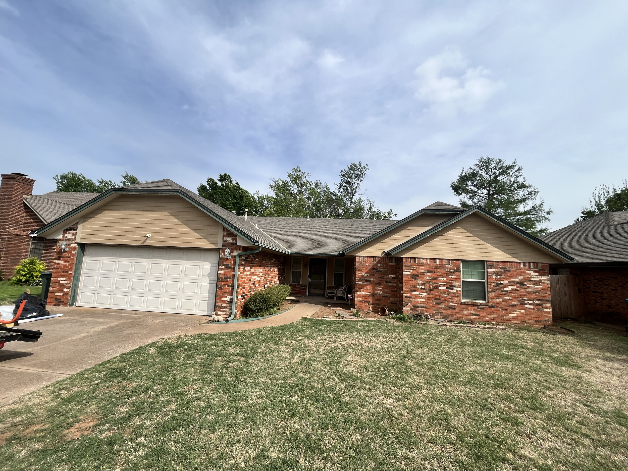 View of a suburban house with a brick and beige siding exterior, a garage door, and a front yard with grass and small bushes. The sky above is mostly cloudy with some blue patches.