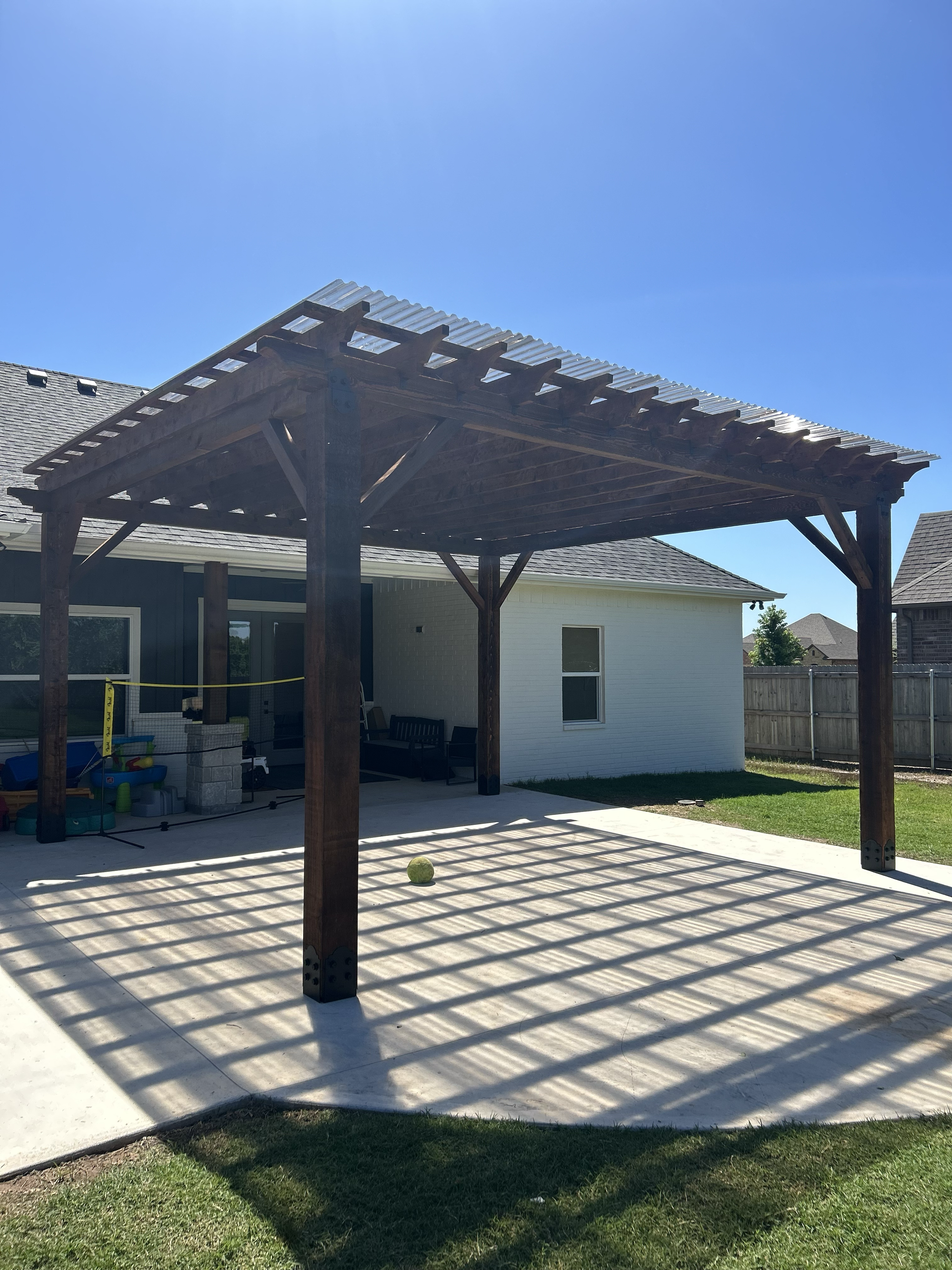 A backyard with a concrete patio, a wooden pergola casting shadows on the ground, a yellow tennis ball, a black bench, a small play area with toys, and a residential house with a white exterior, windows, and a fenced yard.