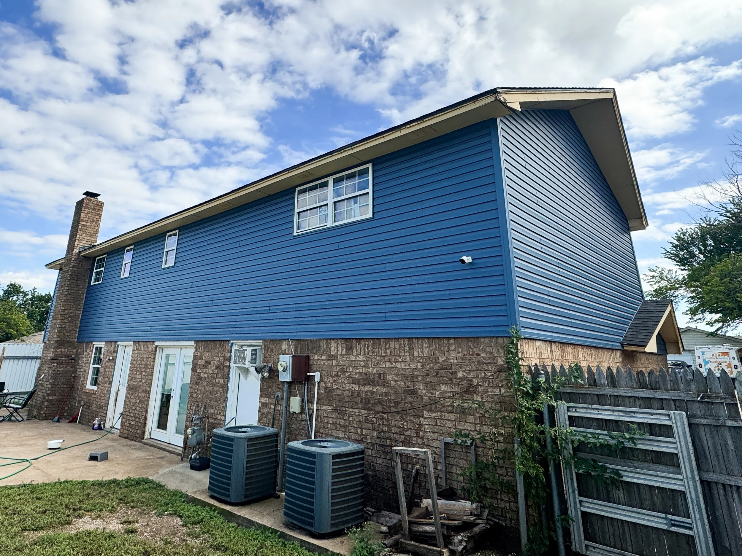 Back view of a two-story house with blue vinyl siding, a brick chimney, multiple windows, a fenced yard, and several air conditioning units outside. The sky is partly cloudy.