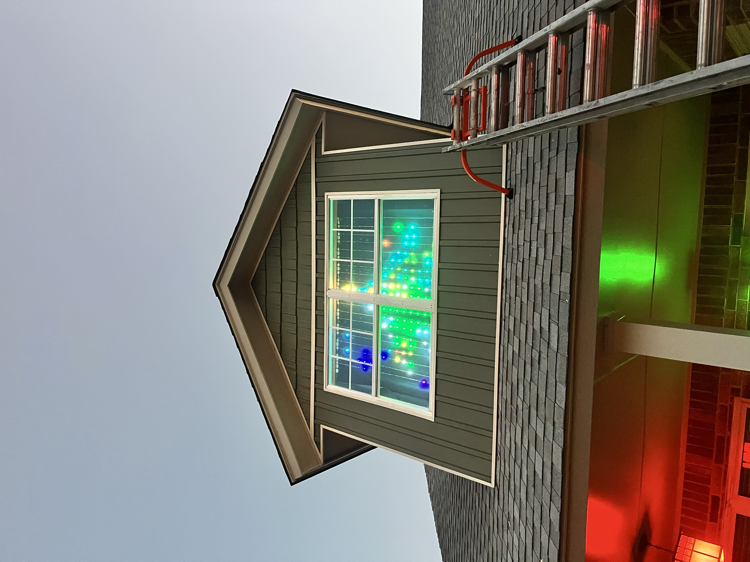 A house with a green exterior and white trim, featuring a large window displaying colorful LED lights creating a pattern; the house has a sloped roof, and a ladder is leaning against the wall. A red bench is visible in the foreground.