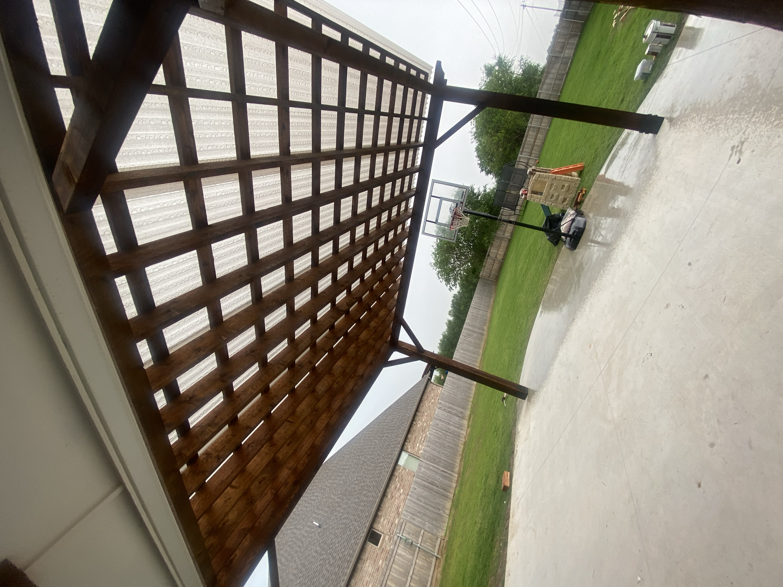 Backyard with a wooden deck, a basketball hoop, green grass, and a fence under an overcast sky.