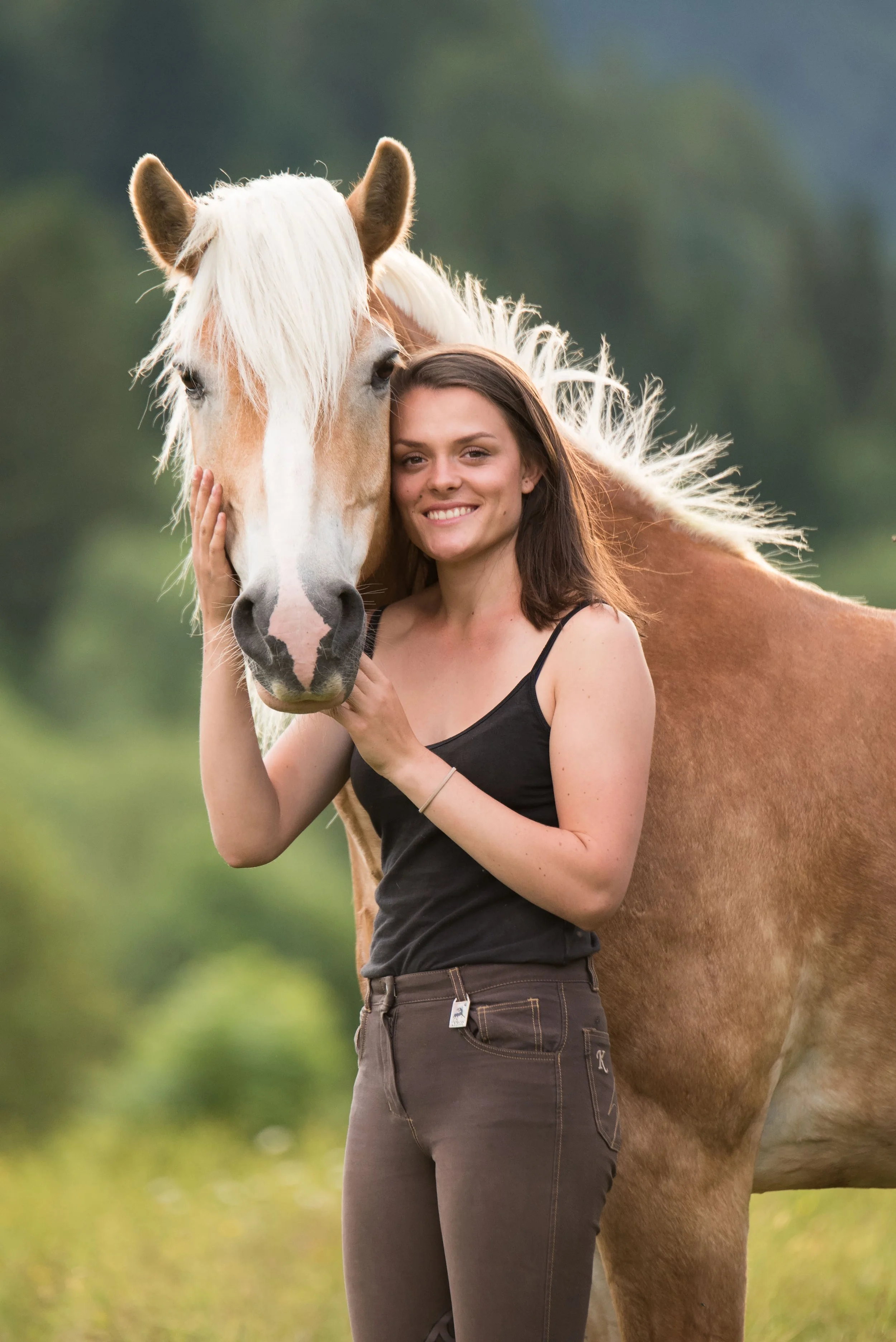 A woman smiling and hugging a light-colored horse with a white mane outdoors with a blurred green background.