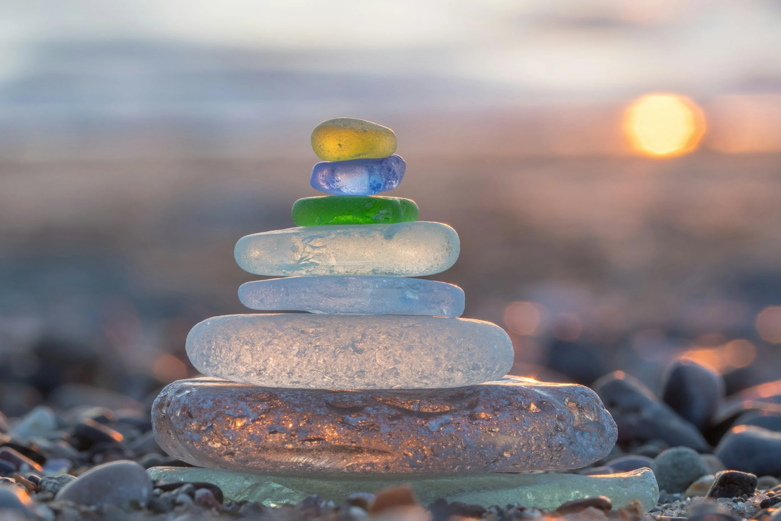 Stack of multi-colored glass stones on a pebble beach at sunset with a blurry horizon and the sun
